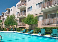 Three-story stucco apartment building with iron balconies overlooking an in-ground pool lined with blue lounge chairs.
